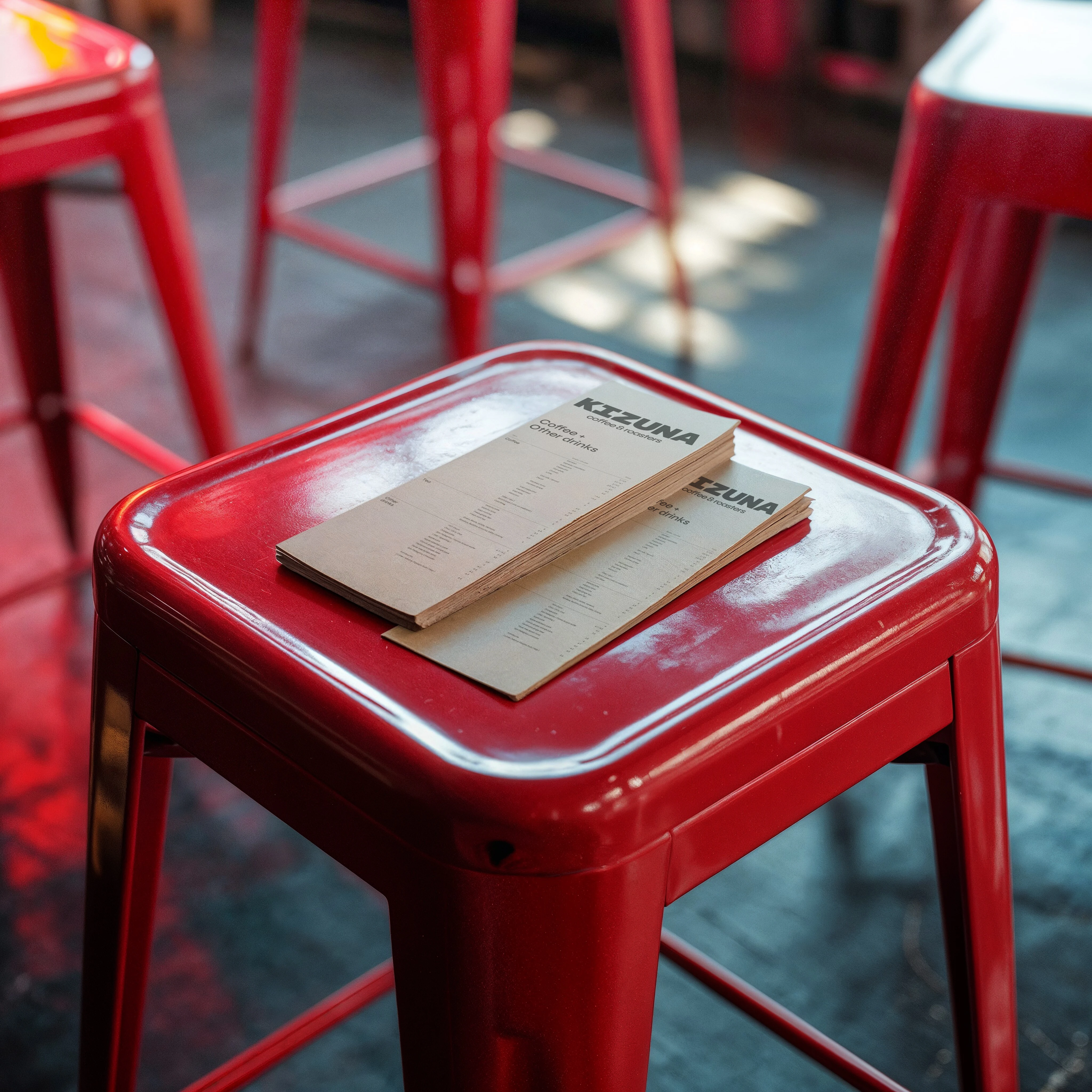 Cartas de bebidas impresas en papel kraft y crema, apiladas sobre un taburete rojo del local